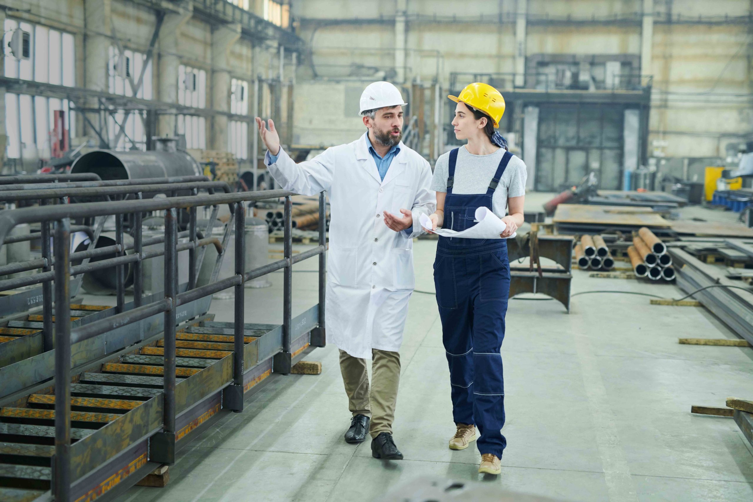 Full length portrait of female factory worker discussing plans with mature engineer while walking  in workshop, copy space